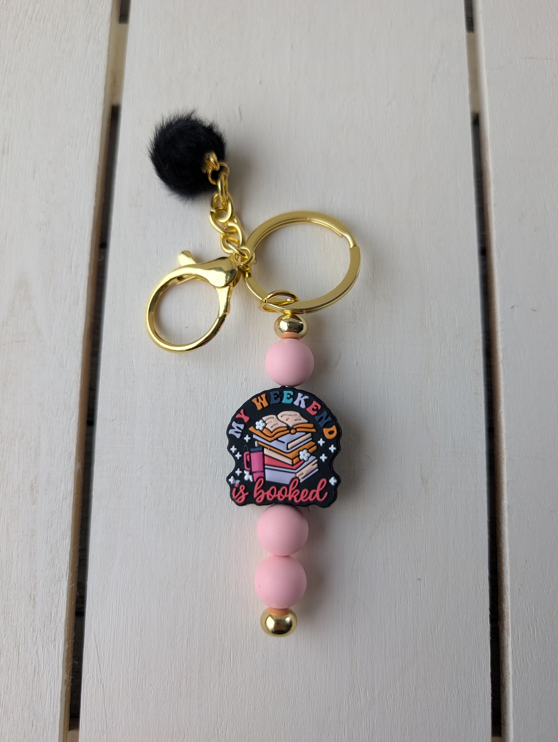 Gold keychain with pink beads and "my weekend is booked" focal bead. Includes black pompom charm.